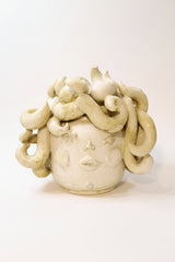 A sculptural ceramic vase with a face-like relief and draped flowing handles, shown on a white surface