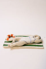 A small sculptural figurine of a reclining figure on a striped green and beige panel with orange accents, photographed against a plain light background.