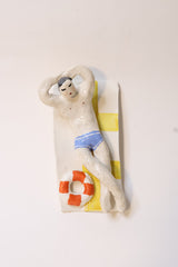A small decorative figurine of a reclining person in a beach-themed setting, with a yellow-and-white striped towel, blue swim trunks, and an orange-and-white life preserver, photographed on a plain light background.
