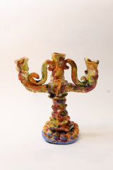 An ornate three-arm candelabra with a sculptural, dragon-like curved design, finished in a multicolored glaze of green, orange, brown, and blue on a white background.