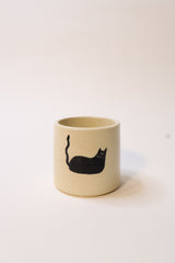 A small cylindrical ceramic cup or tumbler in a warm cream color, featuring a simple black cat silhouette printed on the side, set against a plain off-white background.