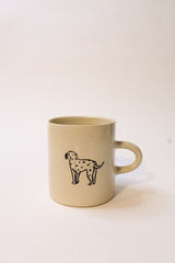 A beige ceramic mug with a handle, featuring a black line-art illustration of a dog with spots, shown against a plain white background.