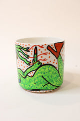 A white ceramic cup featuring a hand-painted green dragon illustration with orange dots and black outlines, set against a plain off-white background.