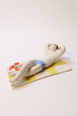A sculptural figurine of a sunbathing figure on a striped towel, with a small orange-red life preserver, photographed against a plain white wall.