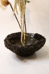 A dark brown, textured ceramic bowl vase holding dried beige and yellow botanical stems, set against a plain off-white background.