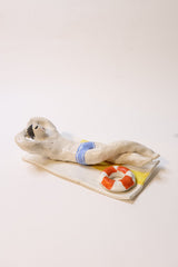 A small sculptural figurine of a person lying on a board or surfboard, with a red-and-white life preserver and blue band detail, photographed against a plain light background.