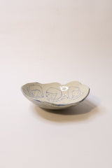 A small handmade ceramic dish with a scalloped rim, featuring blue hand-painted cartoon faces on a cream-colored glaze, set against a plain off-white background.