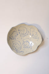 An irregularly shaped cream-colored ceramic dish with hand-drawn blue line illustrations of faces and handwritten text, photographed from above on a plain white background.
